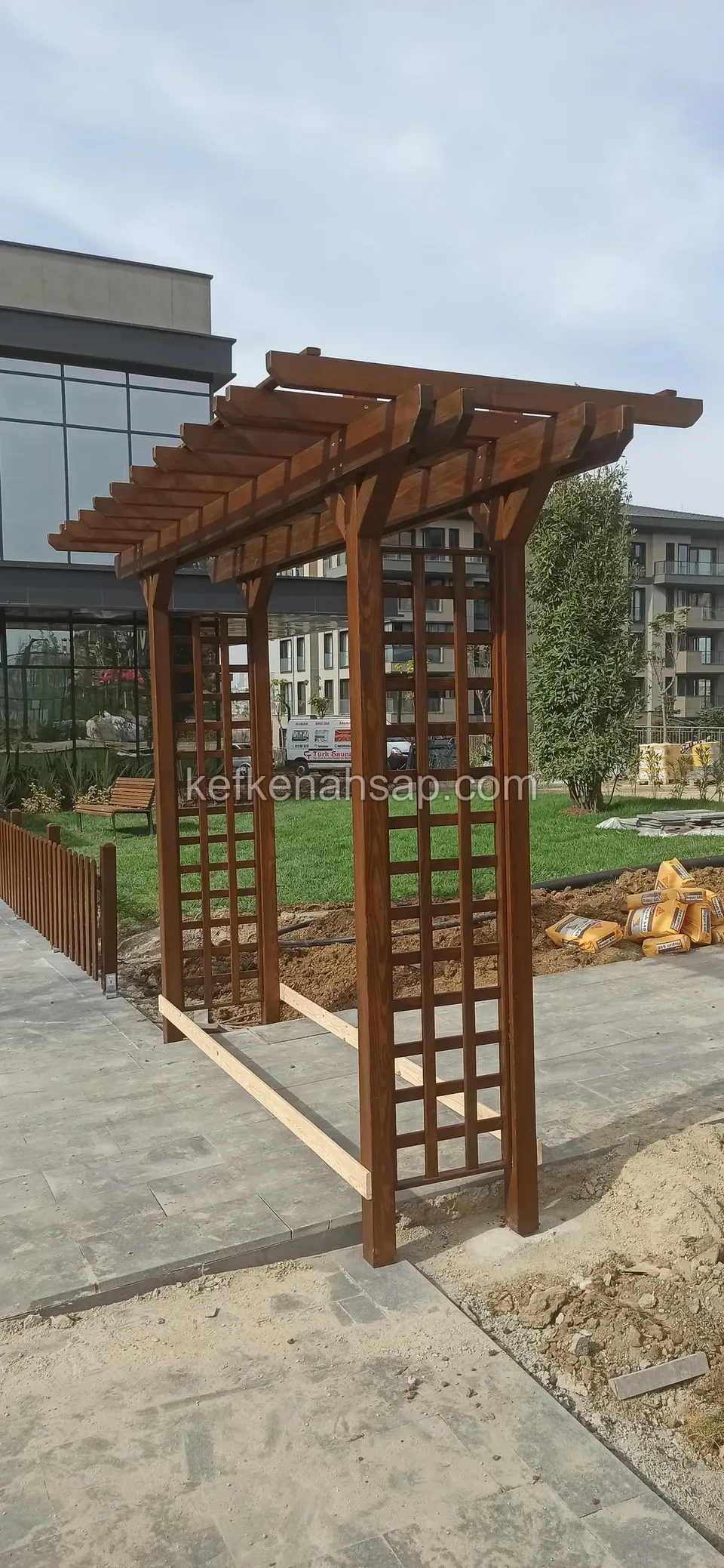 Kafes yan panelli koyu vernikli ahşap pergola kemeri, çevrede kum yığını, taş döşeli yol ve cam cepheli bina