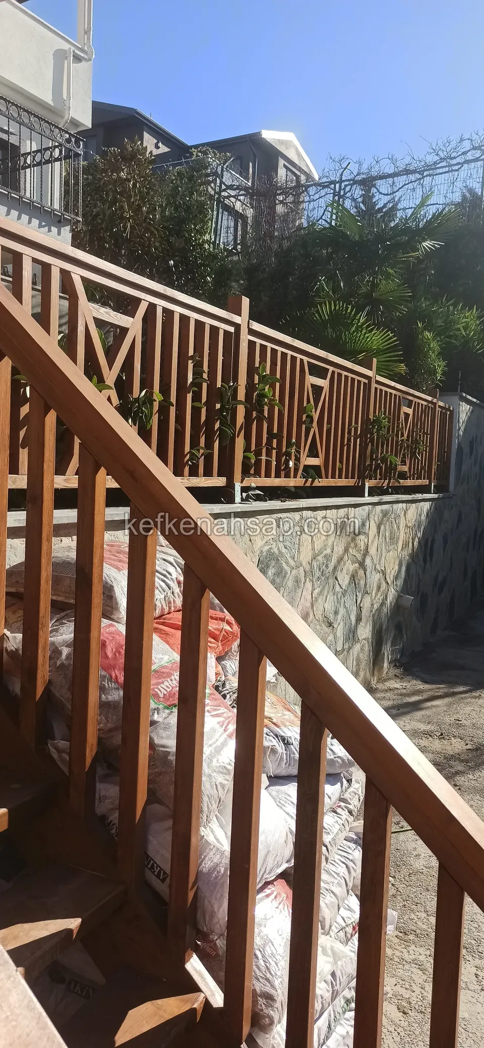 Merdiven boyunca inen kahverengi cilalı ahşap korkuluk, aşağıda taş istinat duvarı üstünde çapraz çıtalı devamı