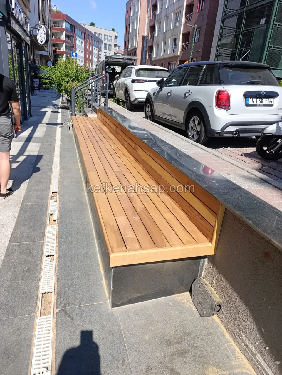 Long wooden bench built against a dark stone wall on a sidewalk, with parked cars and shopfronts beside it