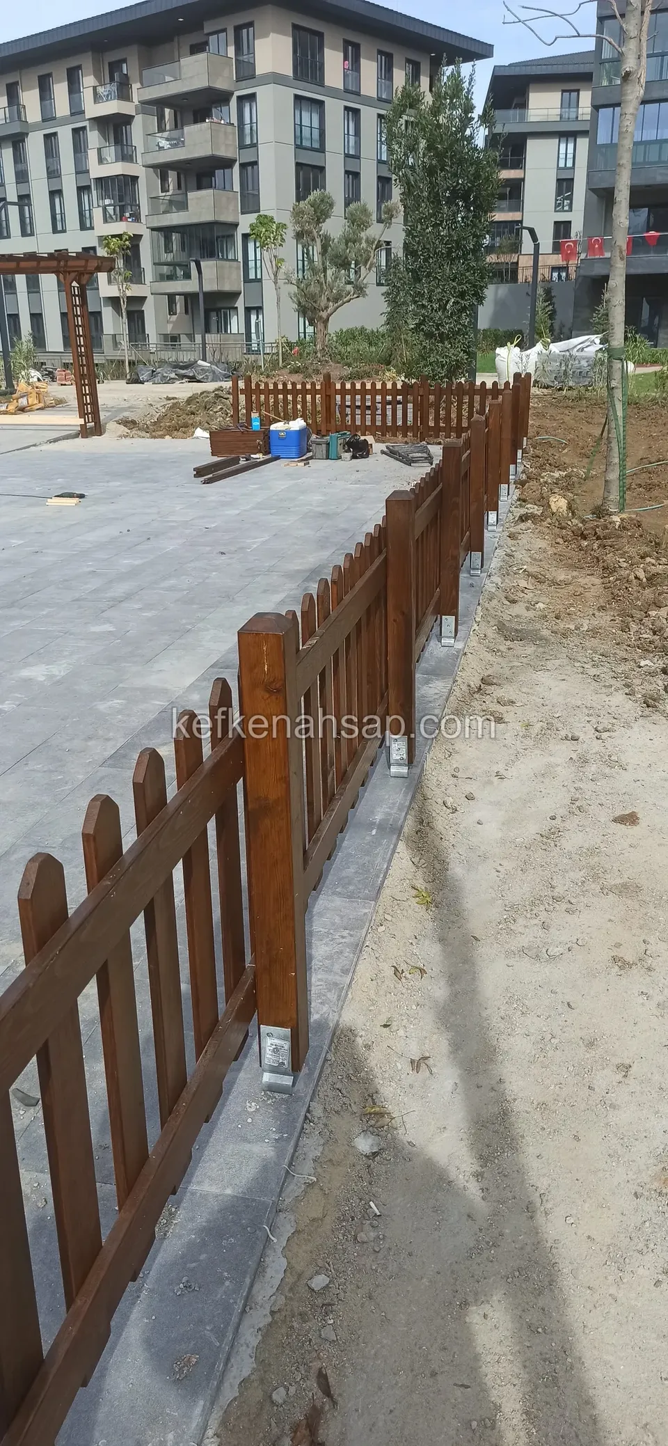 Brown varnished slatted fence lined up on the left of a stone-paved path, with apartment blocks