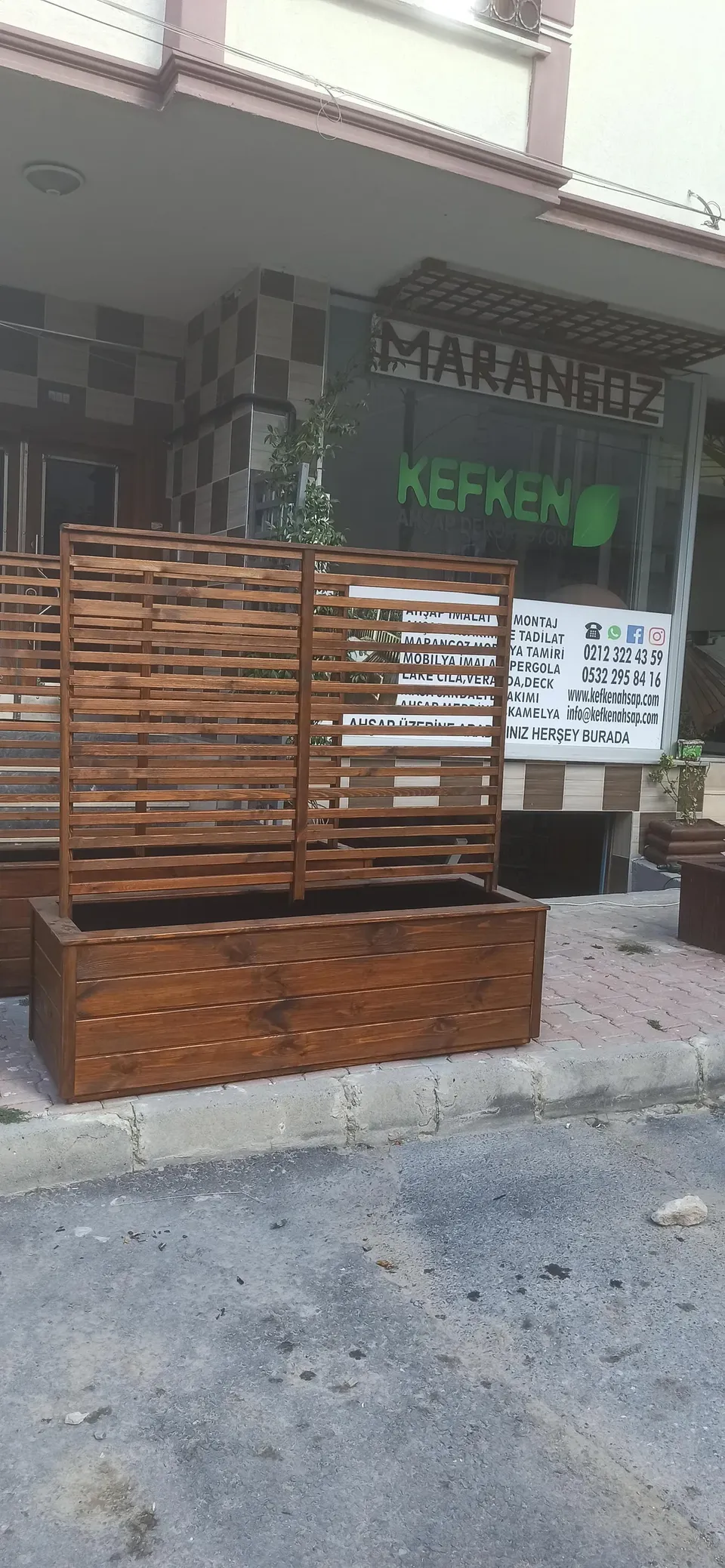 Yol kenarında, kaldırım taşının üstünde duran yatay çıtalı paravanlı ahşap çiçeklik; arkada dükkan cephesi