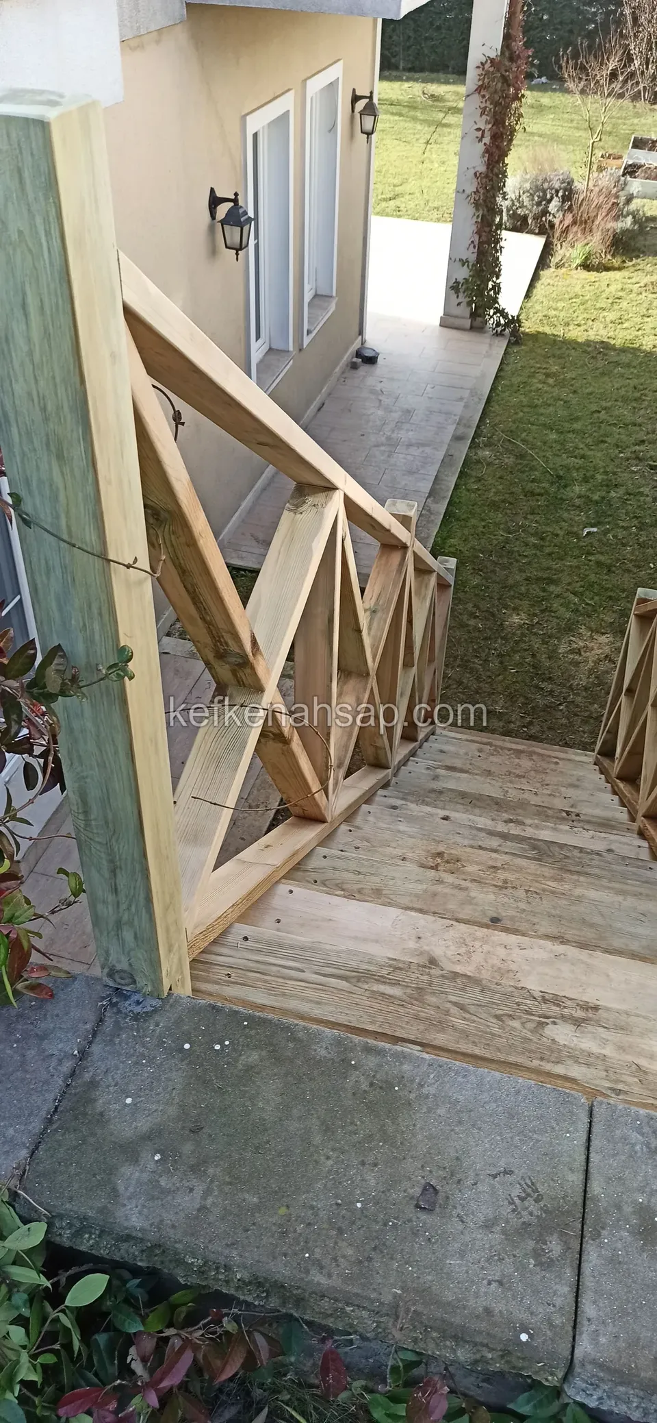 Kreuzstreben-Geländer an kräftigem Pfosten und Treppe mit Bretterstufen neben einem Haus, dahinter eine Fassade mit Glastüren