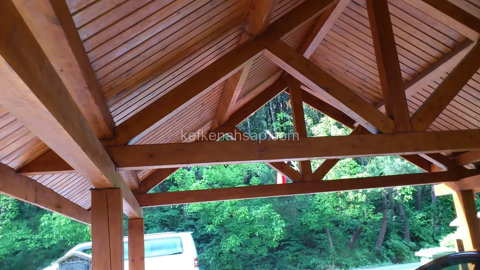Lambri kaplı kamelya çatısının altında kızıl kahve tonlu ahşap makaslar ve arkada yeşil ağaçlar