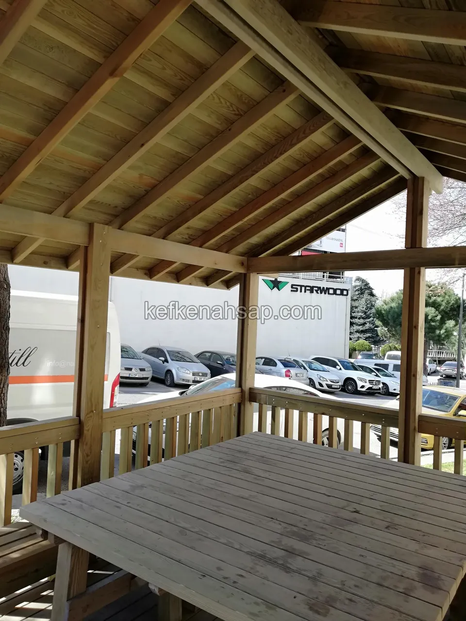 Kamelyanın içinde ahşap masa, çıtalı korkuluk ve çatı kirişleri, dışarıda otopark ve araçlar