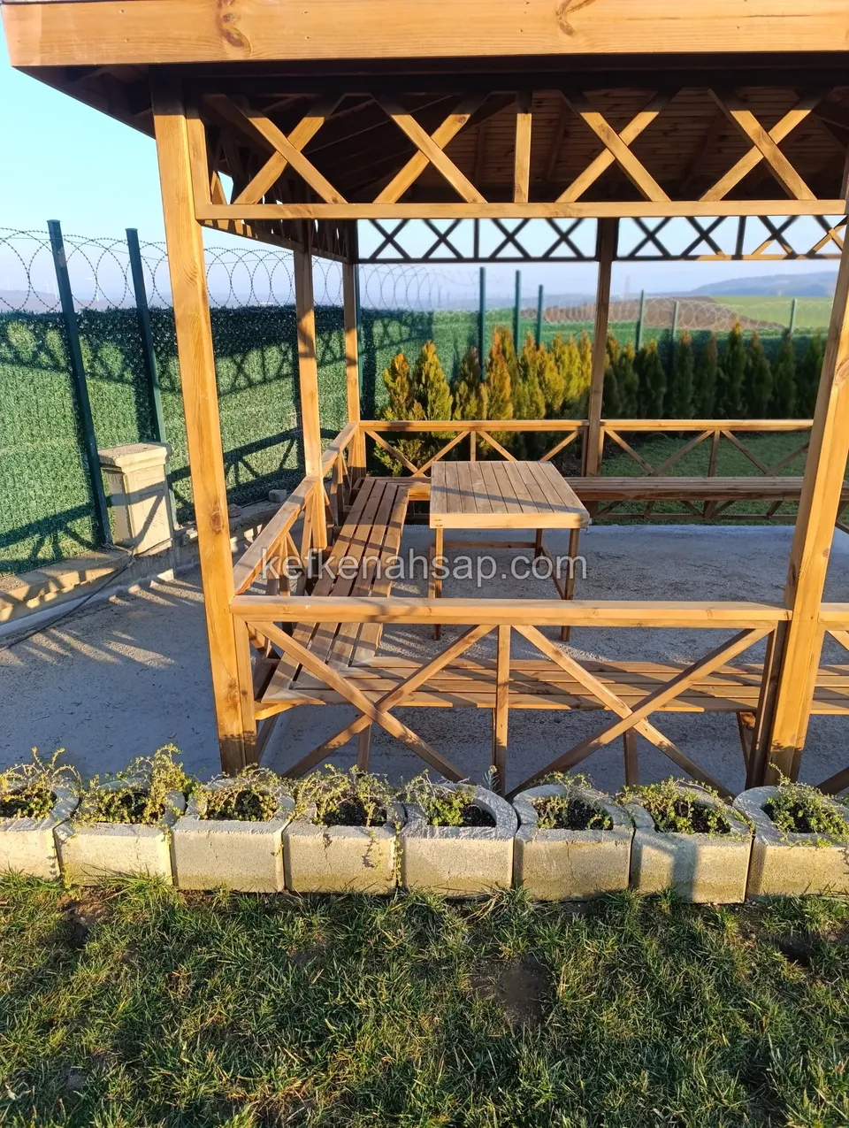 Kamelyanın içinden çapraz çıtalı korkuluk, ahşap masa ve bankların bir kısmı, dışarıda beton çiçeklik blokları
