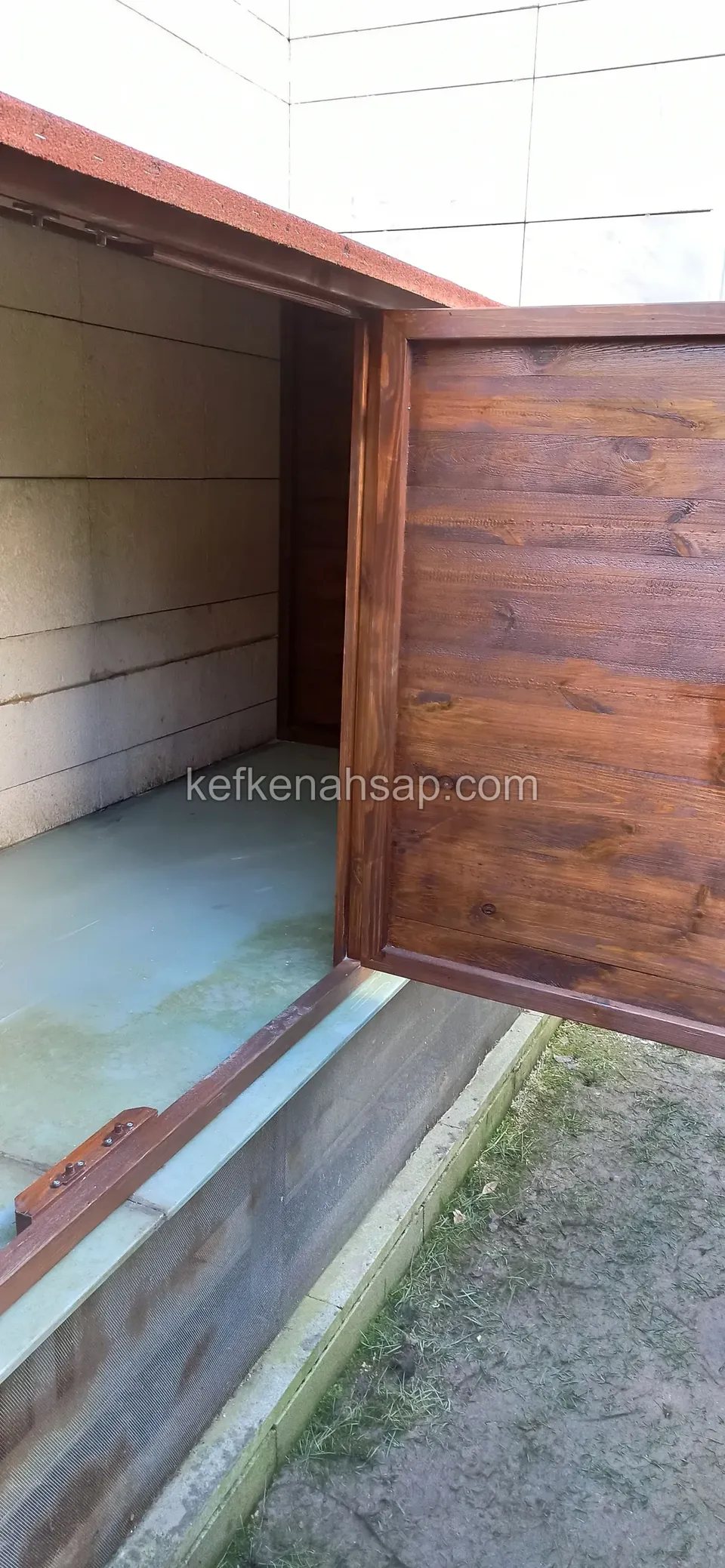 Çıta kaplı ahşap kapağı açılmış, içi boş bir dış mekân dolabı; kırmızı çatılı gövde bir duvara yaslı