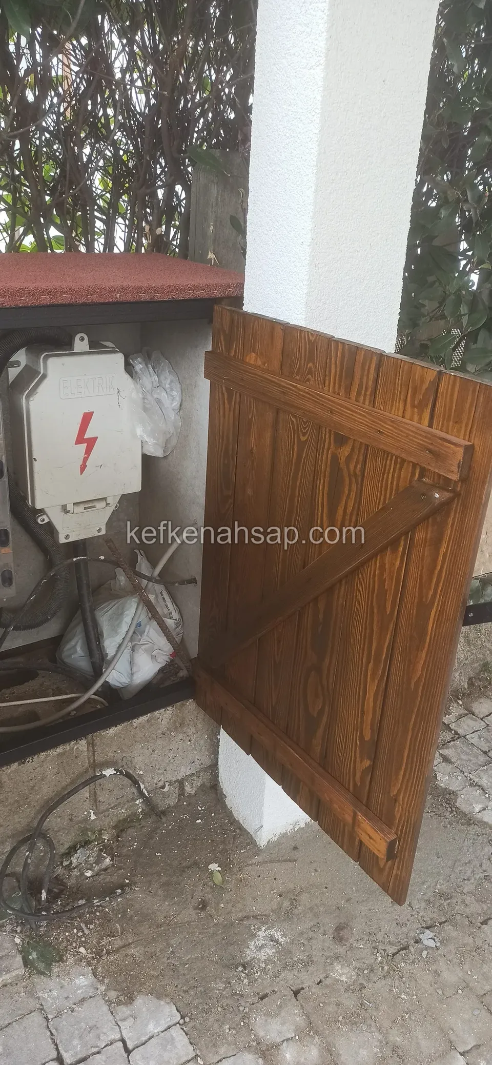 Beyaz kolonun yanında, açık duran Z çaprazlı ahşap dolap kapağı ve içinde şimşek işaretli beyaz bir elektrik panosu