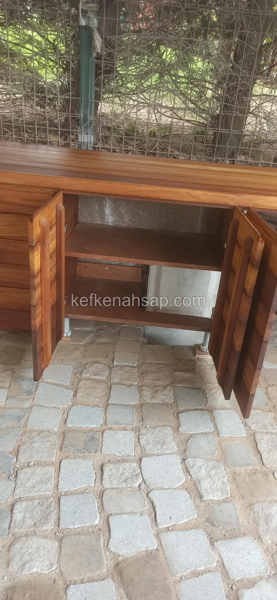 Parke taşlı zeminde, tezgâh altındaki ahşap dolabın iki kapağı açık, ortada tek bir raf bulunan bölme ve çit önünde duruyor