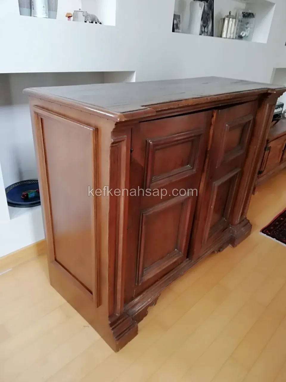 Beyaz nişli bir duvarın önünde, çerçeveli iki kapaklı, koyu kahverengi cilalı ahşap konsol dolabı
