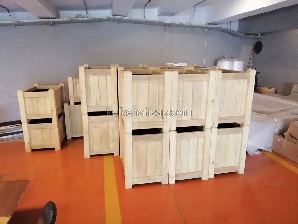 Turuncu zeminli kapalı depoda iki sıra hâlinde yığılmış açık renk ahşap çiçeklik kutuları