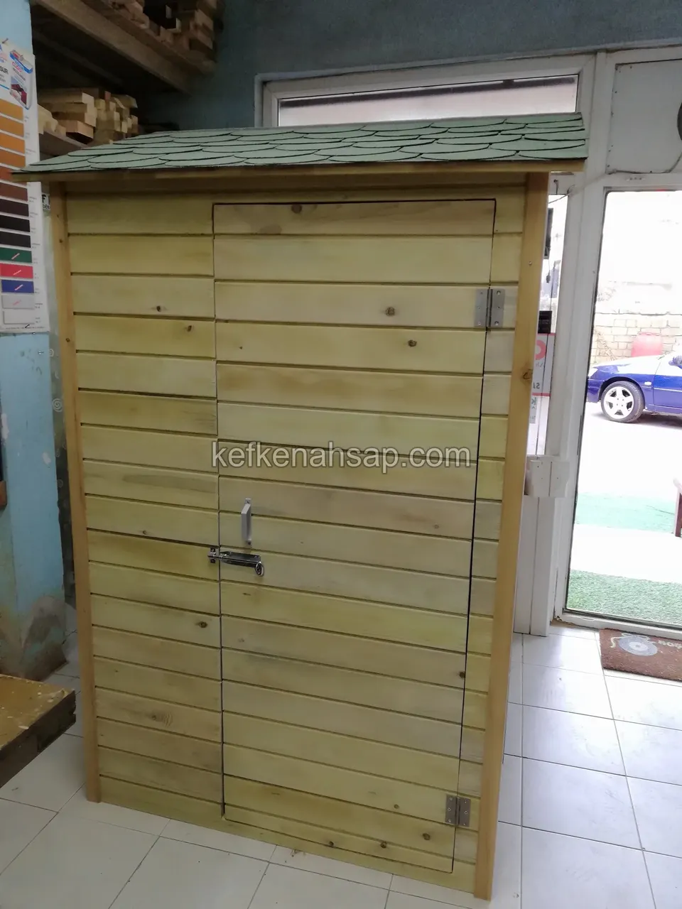 Yatay tahtalarla kaplı, yeşil shingle çatılı, kapağı kapalı ahşap alet dolabı; kapakta metal sürgü var
