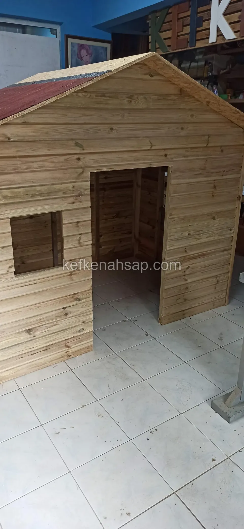 Atölye zemininde duran, kapı ve pencere açıklığı olan, kırmızı çatılı küçük ahşap oyun evi