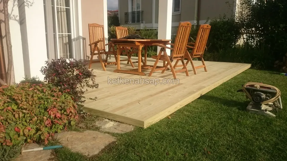 Evin girişinde çimden yükselen açık renkli deck platformu, üstünde ahşap masa ve katlanır sandalyeler, yanında taş basamaklar