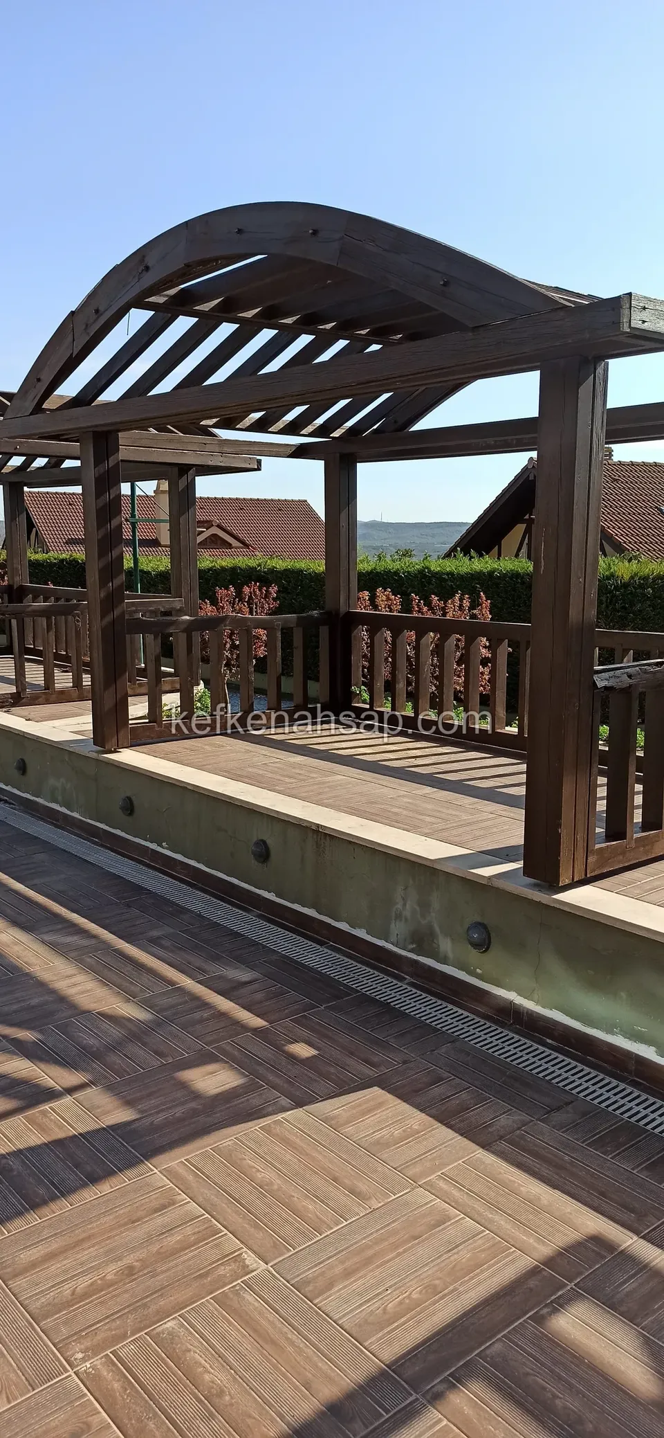 Ahşap desenli zeminli terasta kavisli kirişli çatısı ve ahşap korkuluklarıyla koyu tonlu pergola