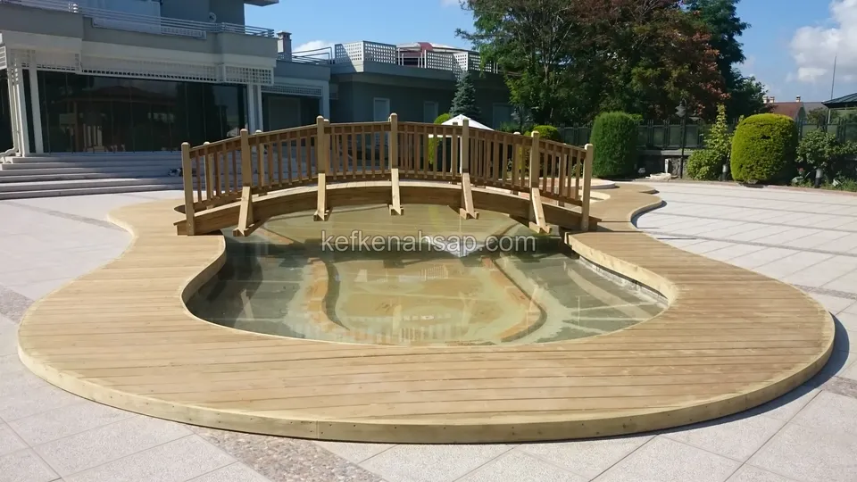 Kavisli sığ havuzu saran açık renkli deck kuşağı ve üstünden geçen kemerli, korkuluklu ahşap köprü, arkada büyük bina