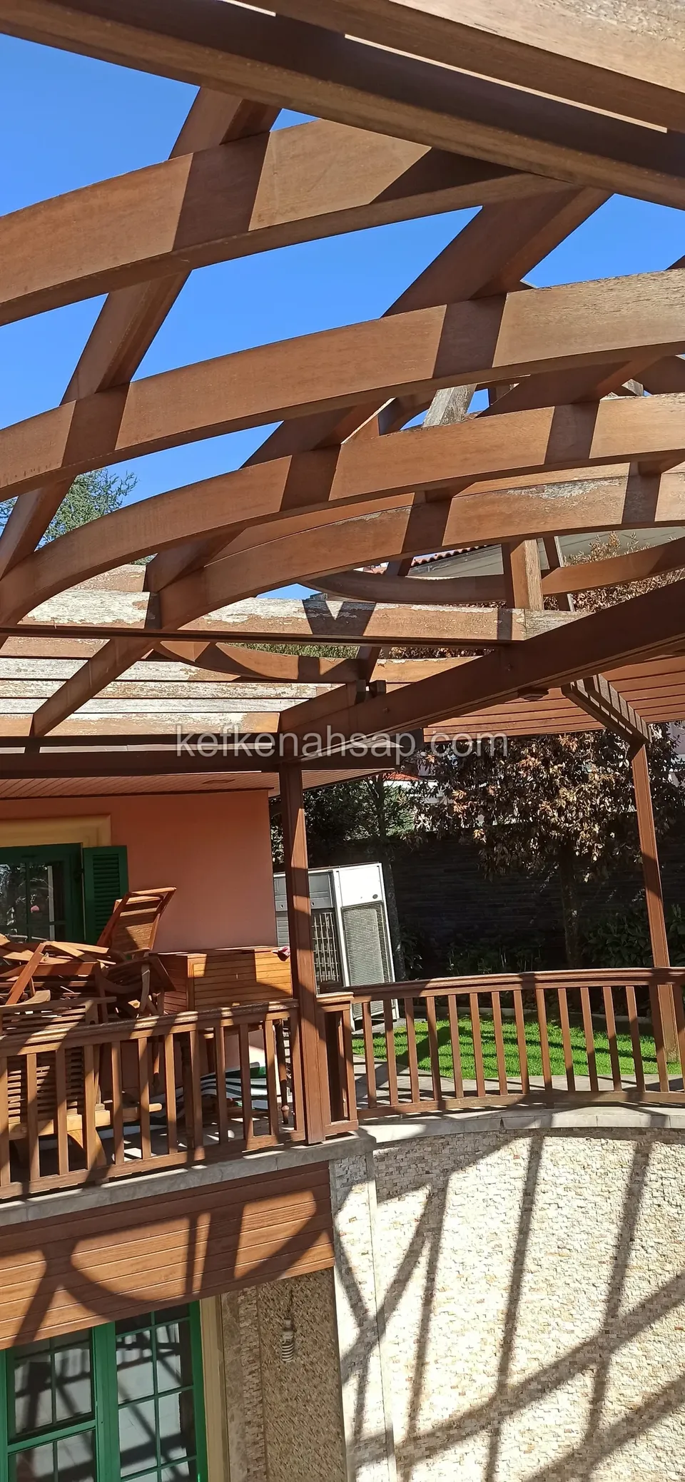 Kavisli pergola kirişleri, ahşap balkon korkuluğu ve yeşil panjurlu pembe renkli bina
