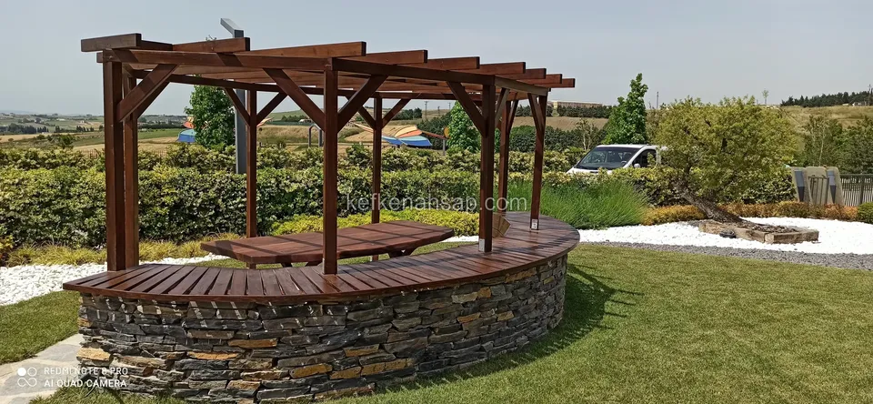Çimenlik bahçede taş örgü kaide üzerinde kavisli ahşap oturak ve üstünde koyu tonlu pergola