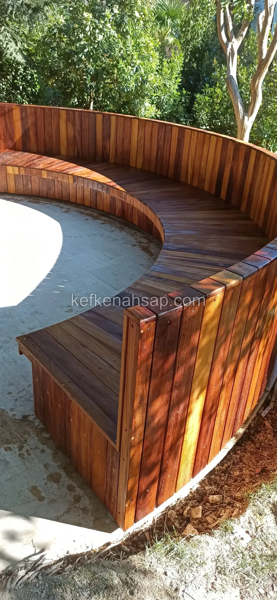 In einem Garten mit Bäumen umschließt eine geschwungene Holzbank mit hoher Lehne eine Betonfläche