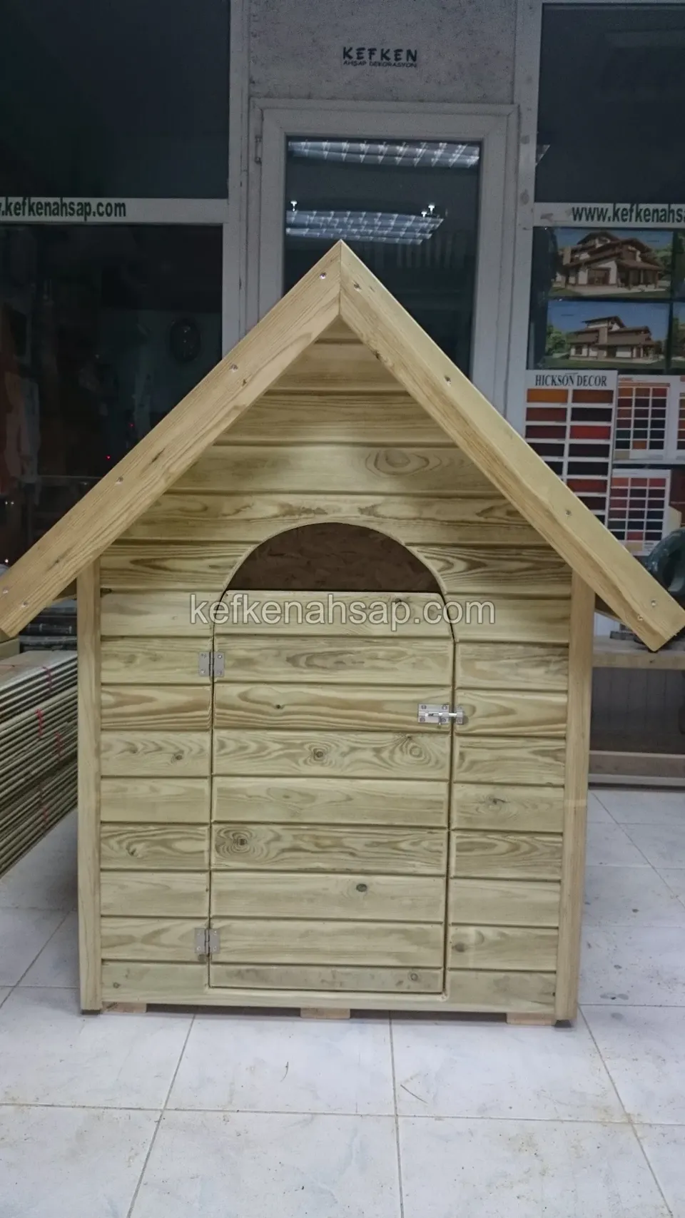 Beşik çatılı, kemer biçimli girişi olan açık renk ahşap köpek kulübesi, karo zeminli bir iç mekânda duruyor