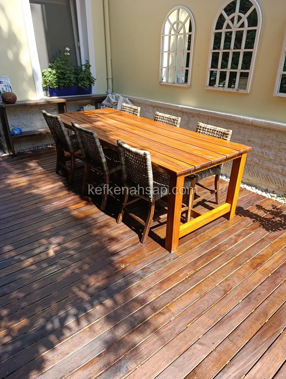 Kemerli pencereli duvarın önünde, koyu tonlu ahşap deck üzerinde ahşap yemek masası ve hasır sandalyeler