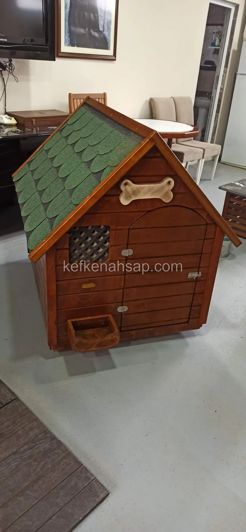 بيت كلب مطلي بالورنيش بسقف أخضر وزخرفة عظمة وفتحة تهوية بشبك وحوض طعام، موضوع في غرفة