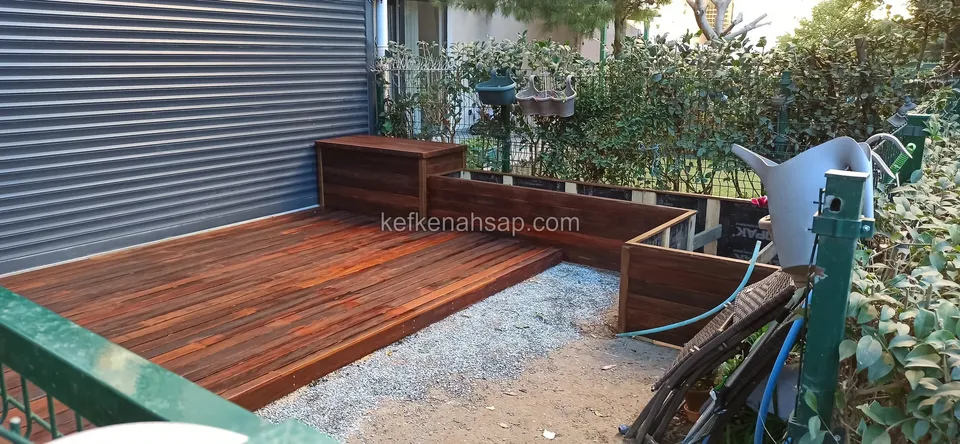 Rotbraunes Deck vor einem Rollladen mit Holzpflanzkästen in der Ecke, einem Kiesstreifen und einer Gießkanne