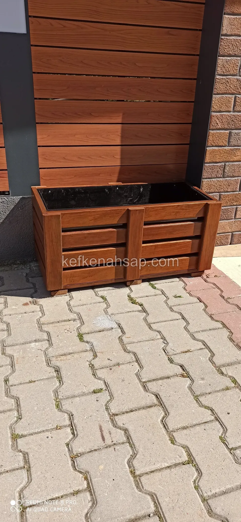 Varnished planter with horizontal slats standing on interlocking paving in front of a timber-clad wall