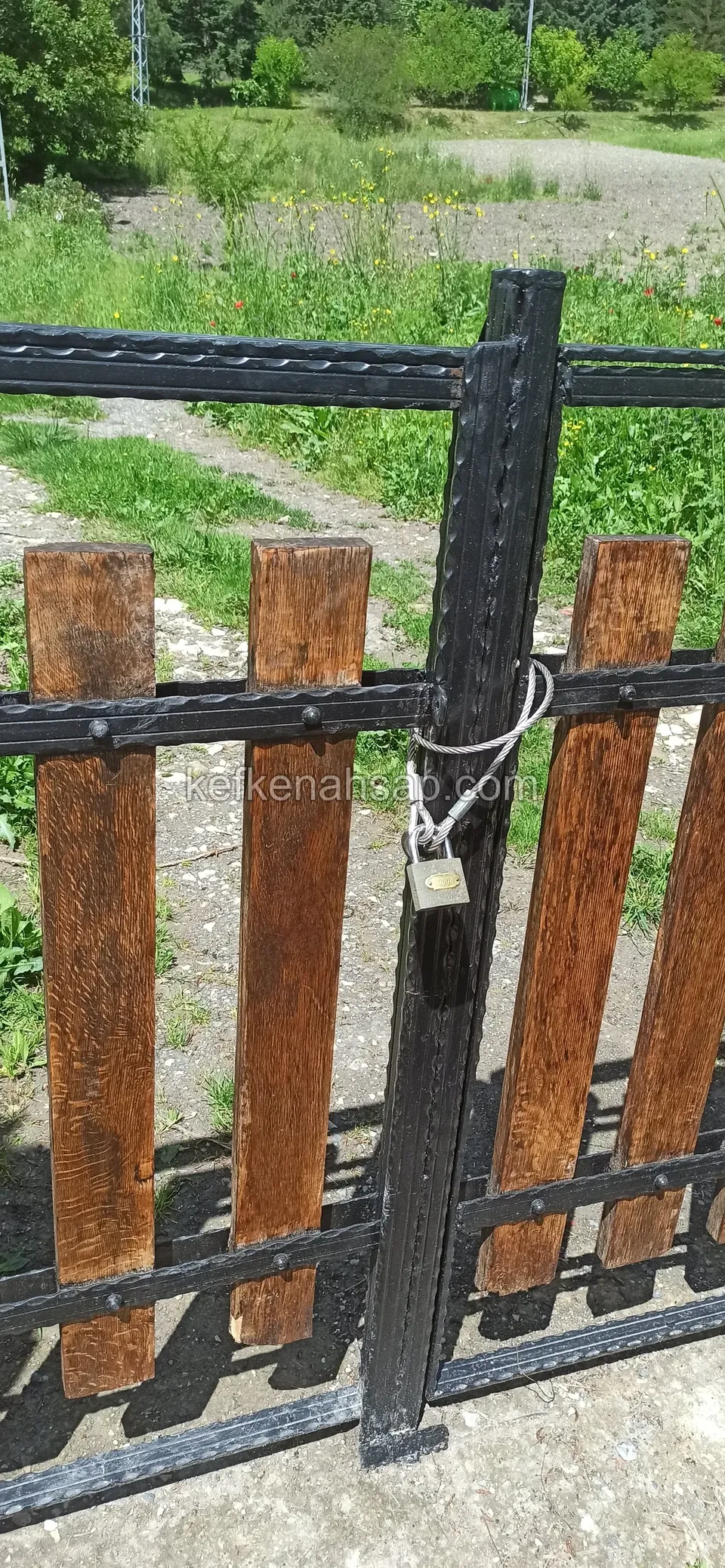 Siyah metal çerçeveli kapının ortasında asma kilitli zincir, yanlarda kahverengi ahşap çıtalar, arkada otlak yol
