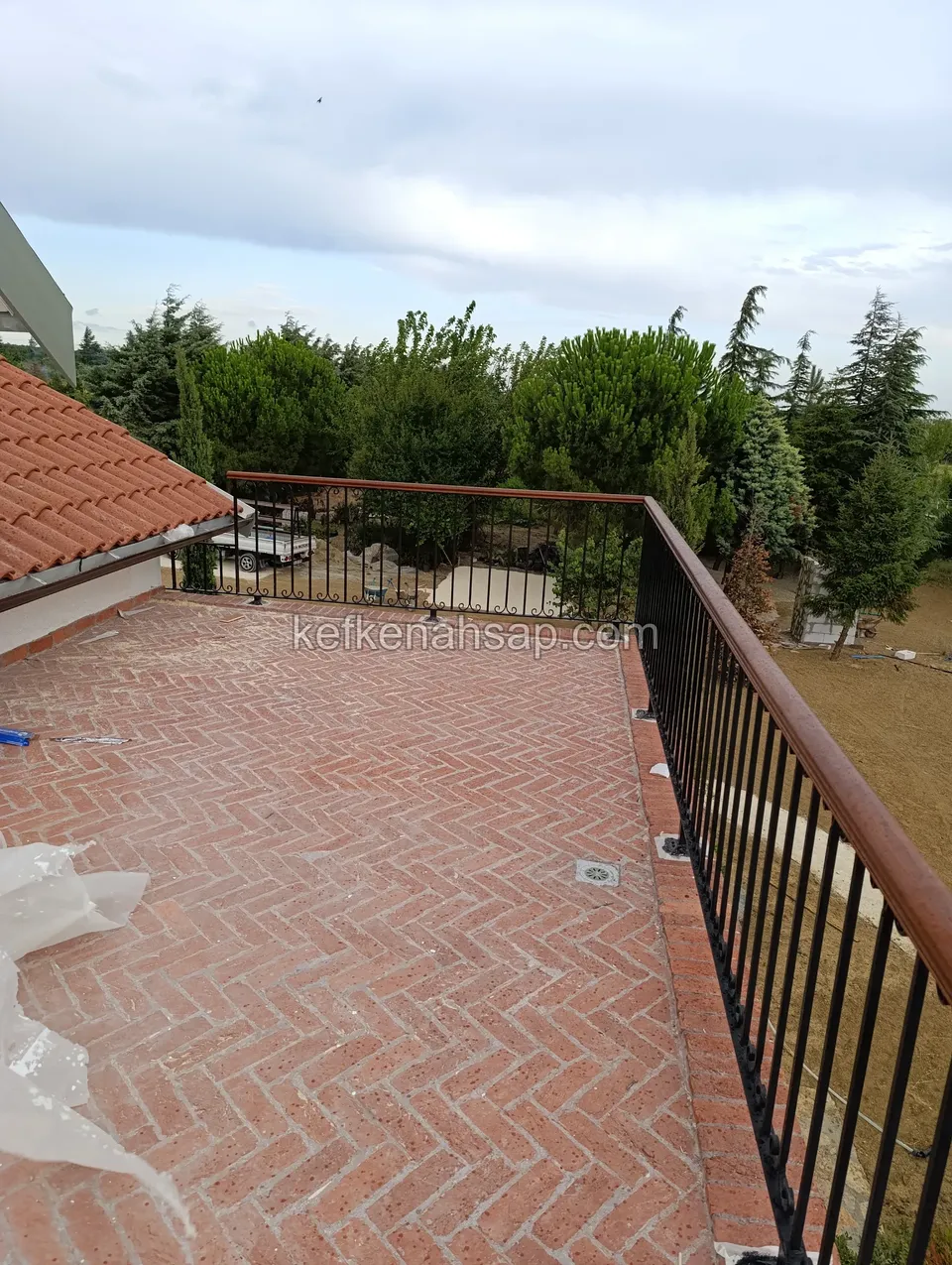 Black vertical-bar iron railing with a timber handrail along a red herringbone brick terrace, with a tiled roof on the left