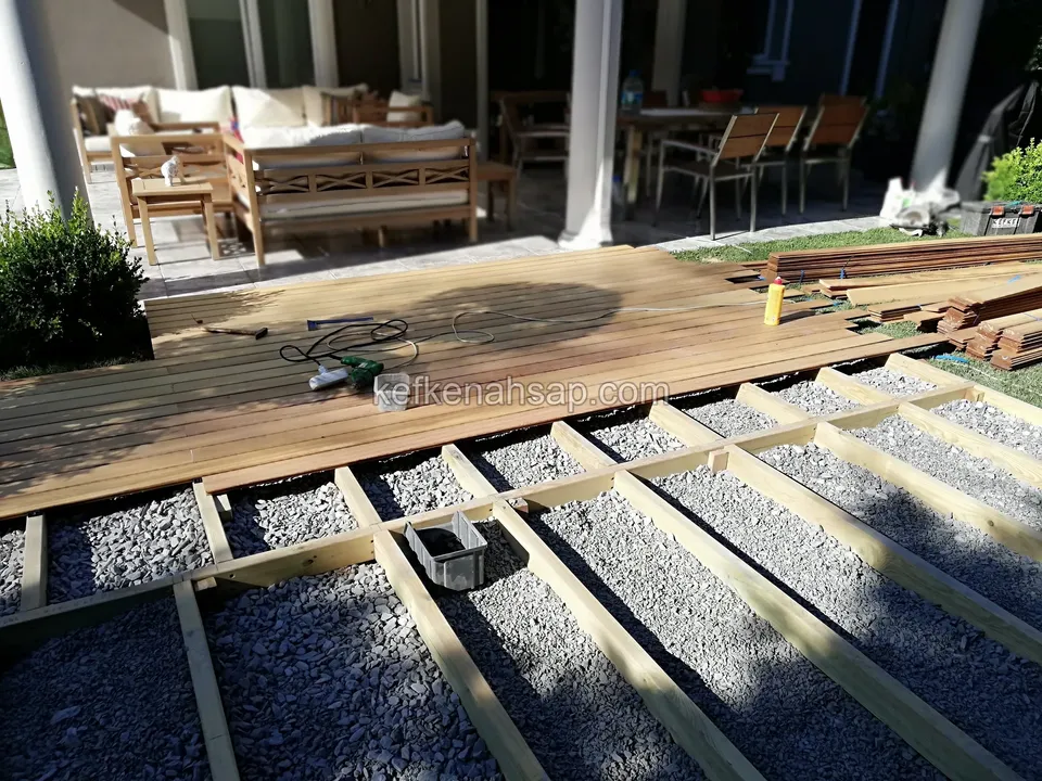 Çakıl üzerinde kiriş iskeleti, bir bölümü deck tahtalarıyla kaplanmış, arkada oturma grubu