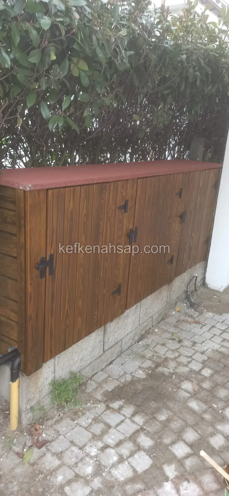 Çalı çitinin dibinde taş tabanlı, koyu bal rengi dikey tahtalı kapaklı, siyah menteşeli, kırmızı çatılı uzun ahşap dolap