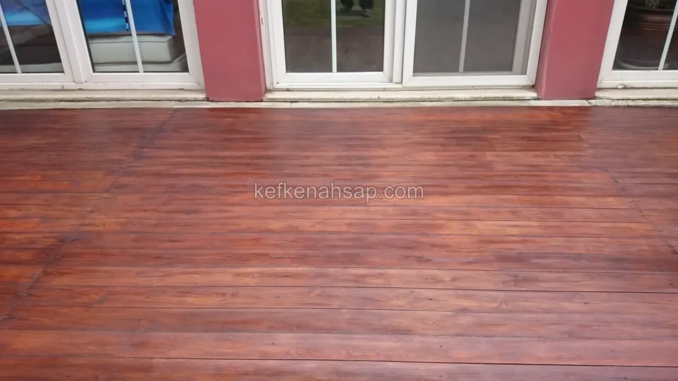 Kırmızı duvarlı evin cam kapıları önünde, kırmızı kahverengiye boyanmış deck; tahtaların damarları boyanın altından seçiliyor