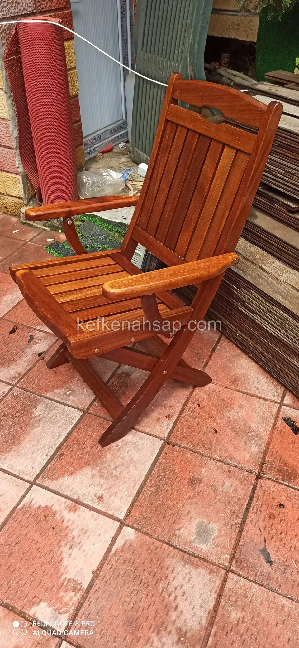 Ein Klappstuhl aus Holz mit Armlehnen und Lattenlehne von der Seite auf einer roten Fliesenterrasse