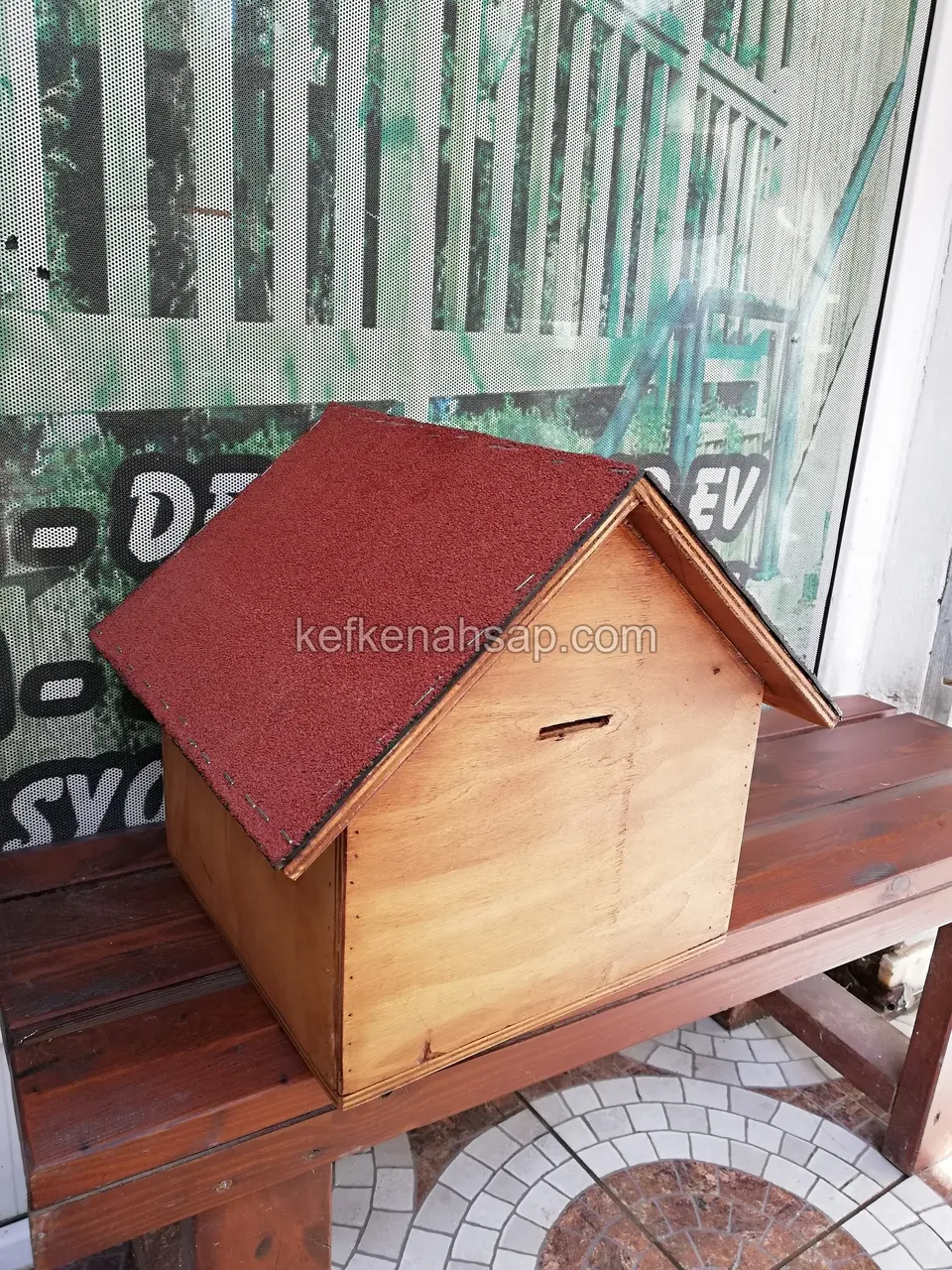 Kırmızı çatılı, vernikli küçük bir ahşap ev; kırmızımsı ahşap bank üzerinde duruyor