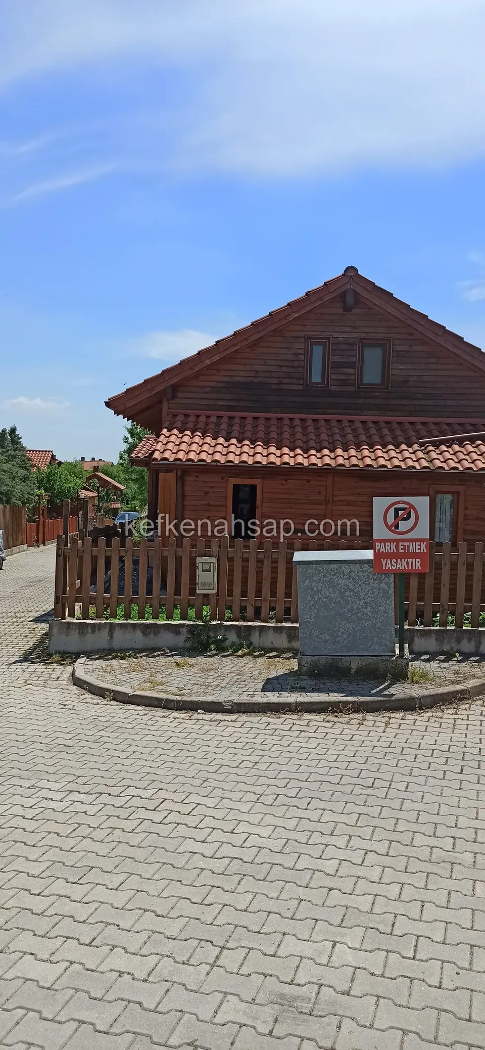 Taş döşeli yolun kenarında, çıtalı ahşap çitle çevrili kiremit çatılı koyu ahşap ev