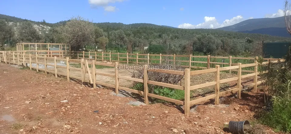 Kırmızı topraklı arazide uzayıp giden iki sıra yatay çıtalı ahşap çit, arkada zeytinlik ve dağ