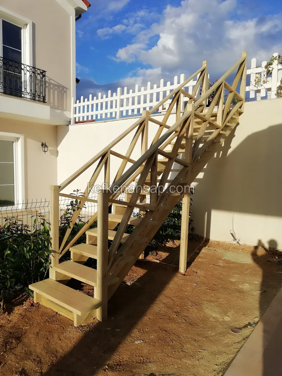 Holztreppe mit Kreuzstreben-Geländer auf rötlicher Erde, oben entlang der Mauer ein weißer Lattenzaun