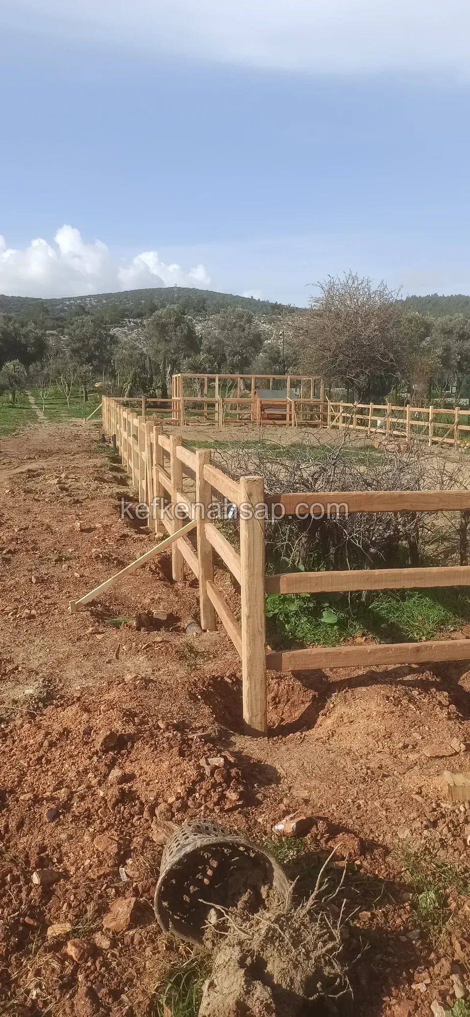 Kırmızı toprak arazide uzun bir dikdörtgeni çeviren yatay çıtalı açık tonlu ahşap çit, önde bir saksı ve kök parçaları