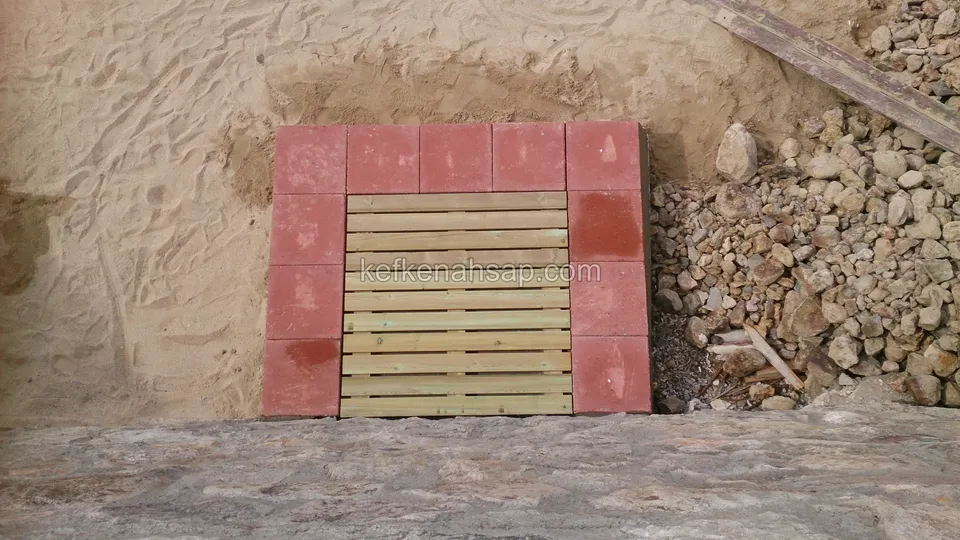 Small horizontally slatted timber vent hatch framed with red paving blocks in a sandy excavation face