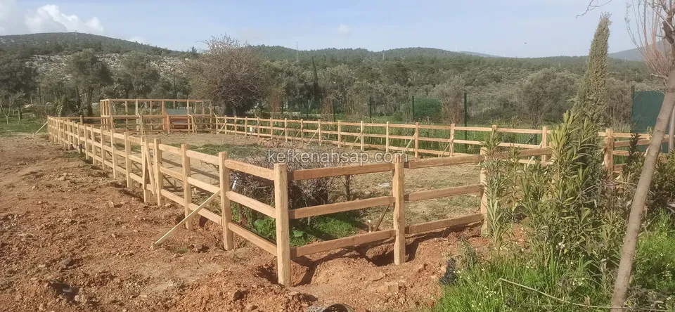 Kırsal arazide yatay kirişli ahşap çitle çevrili geniş bir alan, arkada ağaçlı tepeler
