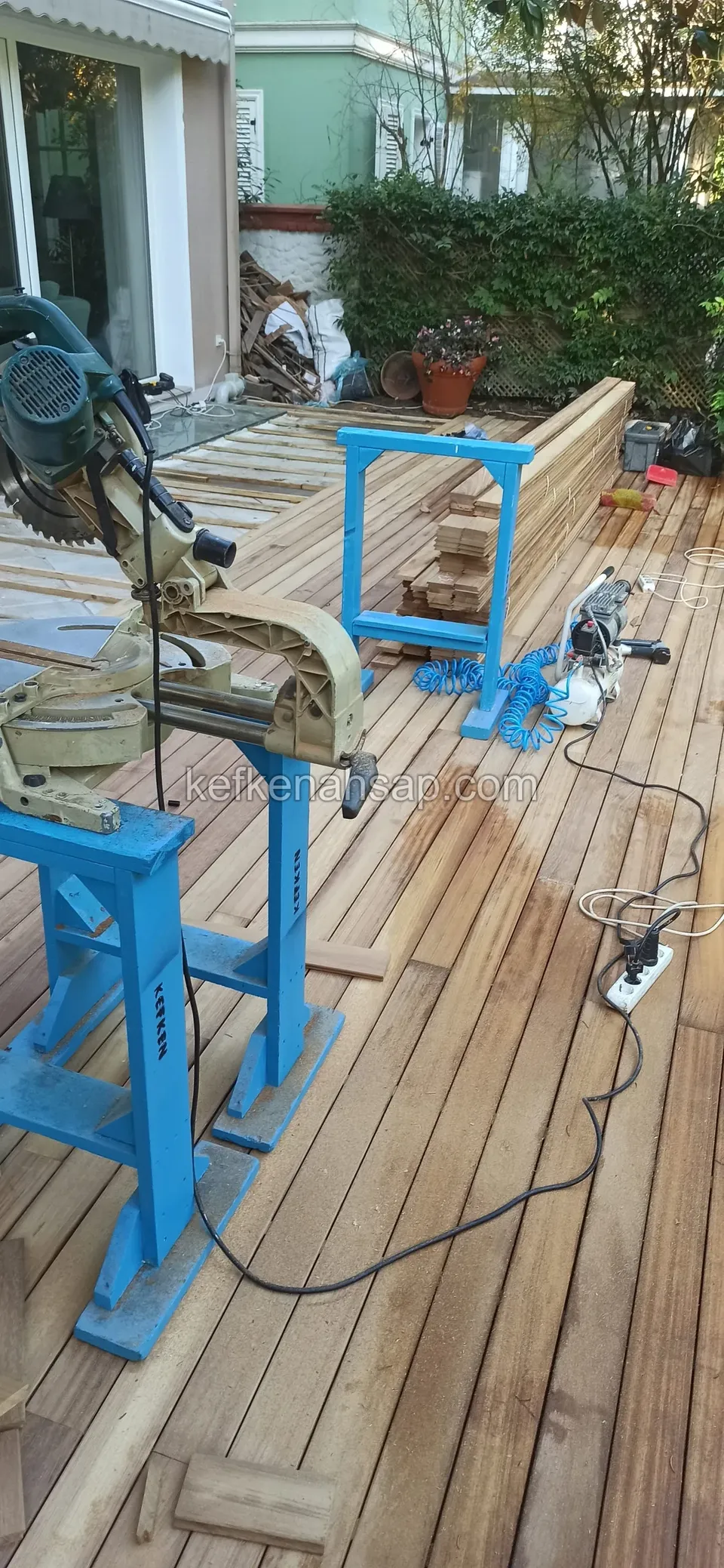 Mitre saw, blue stands and power cables on a part-laid deck, with a glass door behind