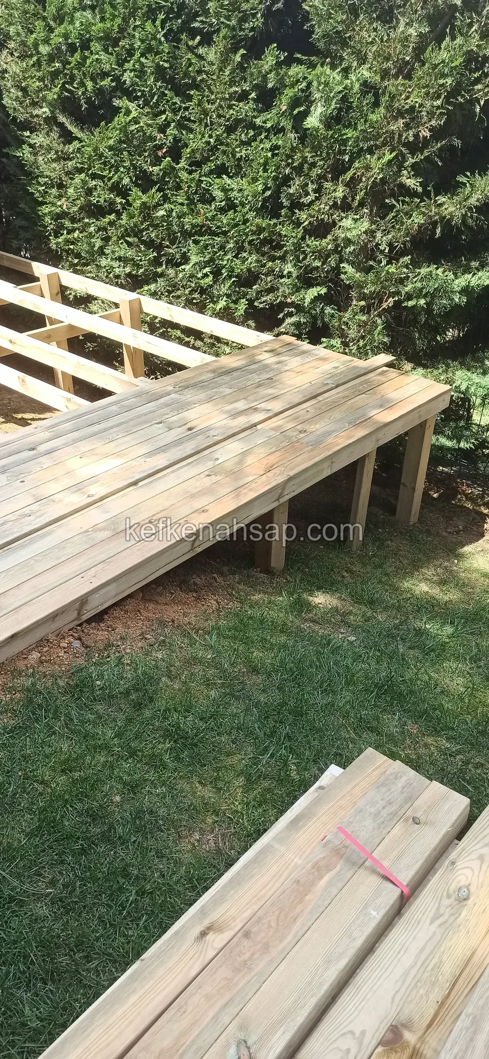 Kısmen tahta kaplı yükseltilmiş deck, önde yığılmış kereste parçaları, arkada yeşil çit