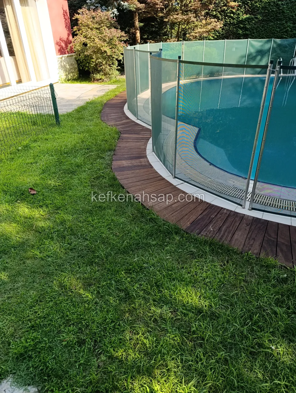 Dark brown deck strip following the curved edge of a pool enclosed by a glass safety fence, beside a lawn
