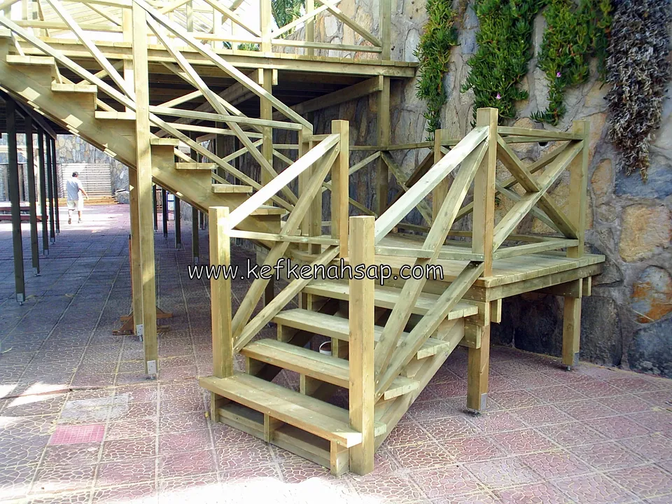 Holztreppe mit Kreuzstreben-Geländer zu einer Plattform nahe dem Meer vor einer Steinmauer, darüber eine Dachkonstruktion