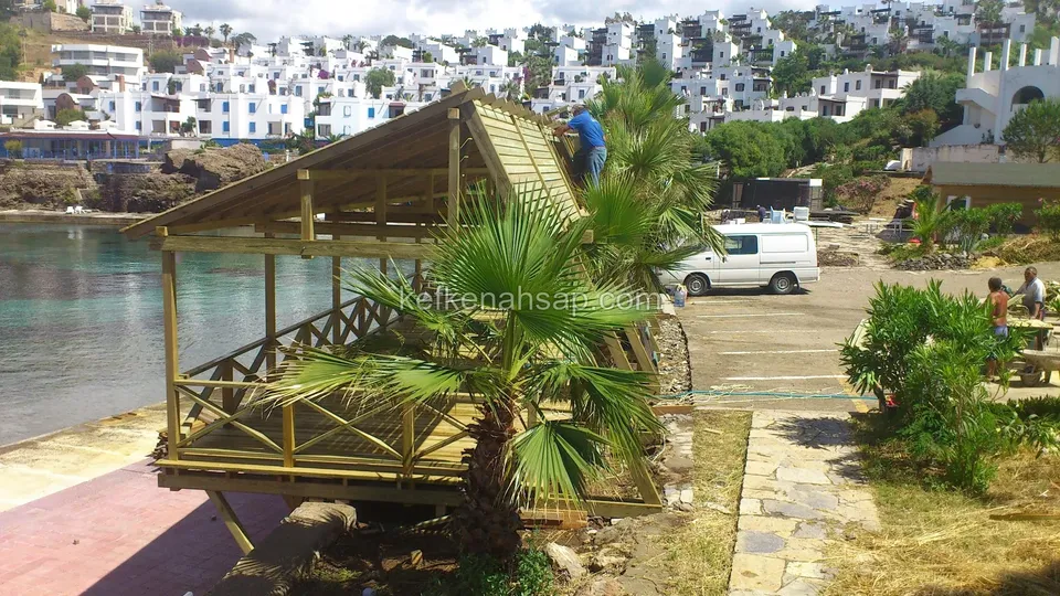 Kıyıda ahşap kamelya, önünde palmiye ve park etmiş beyaz bir araç; arkada körfez ve beyaz evler