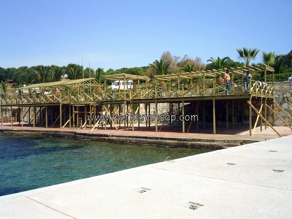 Deniz kıyısında uzun ahşap platformun üzerinde pergola taşıyıcıları ve çalışan işçiler; arkada palmiyeler
