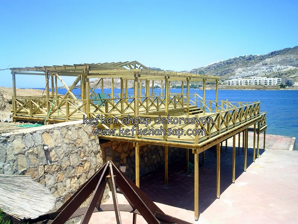 Deniz kıyısında taş duvarın üstünde ayaklarla yükselen, X korkuluklu ve pergola iskeletli deck, uzakta dağlar