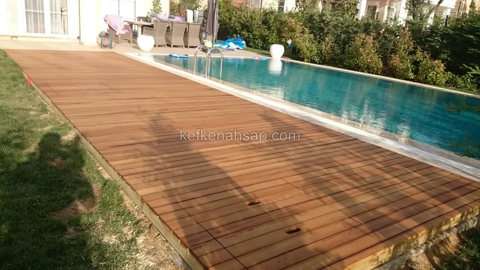 Havuzun yanında uzanan kızıl kahverengi deck, solda çim, arkada bahçe mobilyaları ve ev
