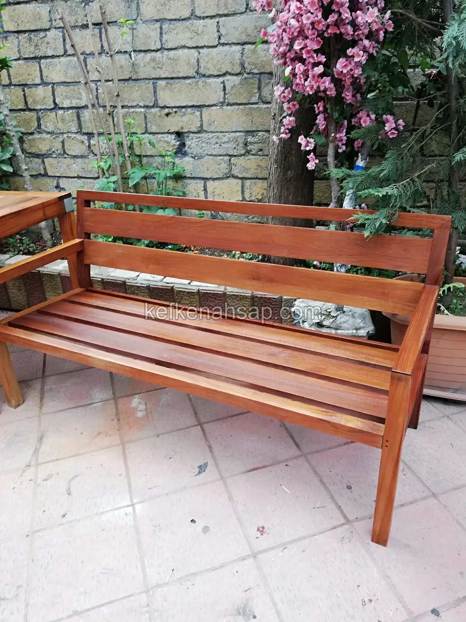 Eine Holzbank mit Armlehnen, Lattensitz und zweiteiliger Rückenlehne vor einer Steinmauer und einem blühenden Baum