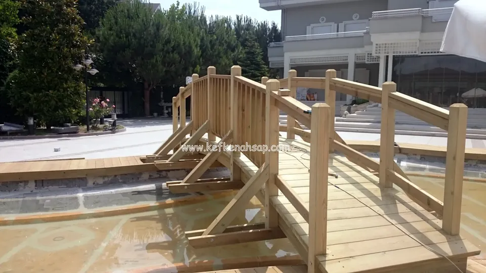 Deck kenarından bakınca köprünün basamağı, kalın dikmeleri ve çapraz destek ayakları; arkada çam ağaçları ve binanın köşesi